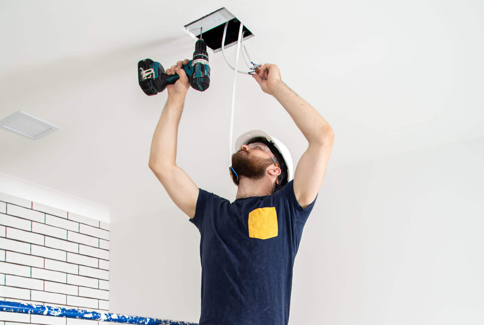Electrician Builder at work, installation of lamps at height. Professional in overalls with a drill. On the background of the repair site.
